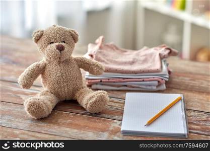 babyhood and clothing concept - baby clothes, teddy bear toy and notebook with pencil on wooden table at home. baby clothes, teddy bear toy and notebook