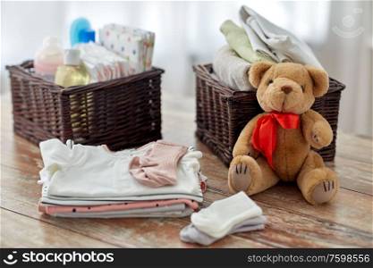 babyhood and clothing concept - baby clothes, teddy bear toy and wicker baskets on wooden table at home. baby clothes and teddy bear toy on table at home