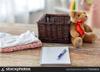 babyhood and clothing concept - baby clothes, teddy bear, toy blocks and notebook with pen on wooden table at home. baby clothes, teddy bear, toy blocks and notebook
