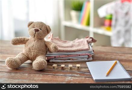 babyhood and clothing concept - baby clothes, teddy bear, toy blocks and notebook with pencil on wooden table at home. baby clothes, teddy bear, toy blocks and notebook
