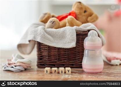 babyhood and clothing concept - teddy bear in wicker basket with baby things and toy blocks on wooden table at home. teddy bear toy in basket with baby things on table
