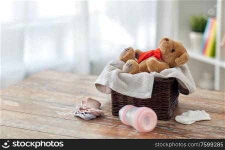 babyhood and clothing concept - teddy bear toy in wicker basket with baby things on wooden table at home. teddy bear toy in basket with baby things on table