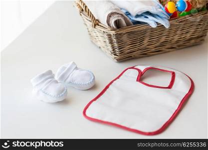 babyhood, childhood, motherhood and object concept - close up of white baby bootees, bib and newborn stuff in basket on table