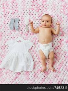 babyhood, clothes and people concept - sweet little baby girl in diaper lying with dress, socks and hairbrush on knitted pink blanket of plush yarn. baby girl in diaper lying with dress on blanket