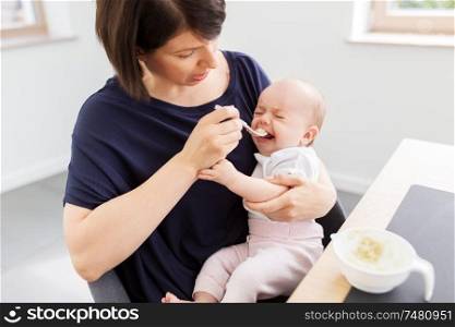 babyhood, motherhood and food concept - middle-aged mother spoon feeding crying baby daughter with puree at home. middle-aged mother feeding baby daughter at home