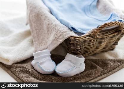 babyhood, motherhood and object concept - close up of white baby bootees with pile of clothes and towel for newborn boy in basket on table