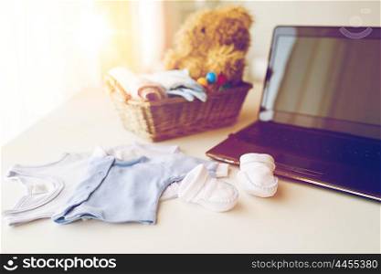 babyhood, motherhood, clothing, technology and object concept - close up of baby clothes and toys for newborn boy in basket with laptop computer at home