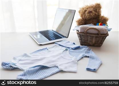babyhood, motherhood, clothing, technology and object concept - close up of baby clothes and toys for newborn boy in basket with laptop computer at home