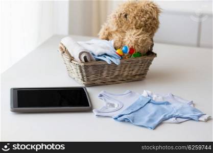 babyhood, motherhood, clothing, technology and object concept - close up of baby clothes and toys for newborn boy in basket with tablet pc computer at home