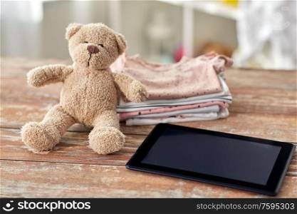 babyhood, technology and clothing concept - baby clothes, teddy bear toy and tablet pc computer on wooden table at home. baby clothes, teddy bear toy and tablet computer