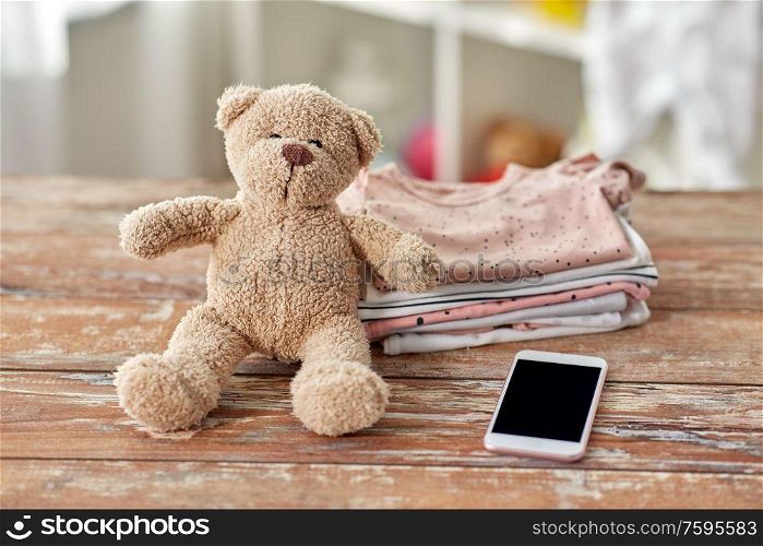 babyhood, technology and clothing concept - baby clothes, teddy bear toy and smartphone on wooden table at home. baby clothes, teddy bear toy and smartphone