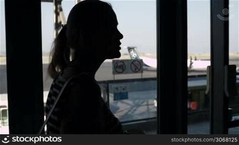 Back-lit silhouette of a young woman drinking coffee at the airport and looking through the window at the airplanes