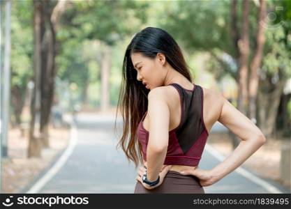 Back of Asian sport woman show action of waist or back pain after exercise in park or garden with day light.