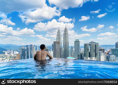 Back of tourist in a swimming pool on rooftop with Kuala Lumpur downtown view and blue sky. Malaysia travel trip in vacation and holidays concept in Asia. Skyscraper and high-rise buildings at noon.