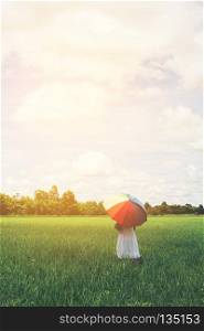 Back Of Young Beautiful woman holding multicolored umbrella in green grassland field and cloud blue sky.. Back Of Young Beautiful woman holding multicolored umbrella in g