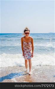 Back rear view of young woman in summer dress standing by the sea or the ocean seaside beach on vacation travel holiday looking to the camera smiling