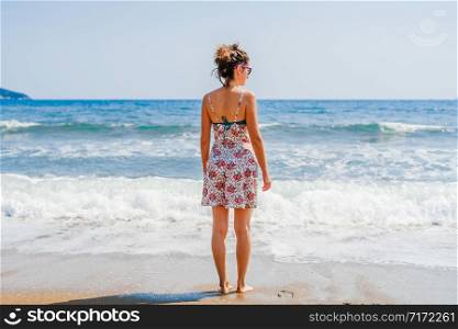 Back rear view of young woman in summer dress standing by the sea or the ocean seaside beach on vacation travel holiday looking to the sea