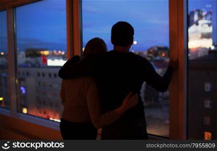 Back shot of young loving couple by the window at night. They hugging and enjoying city view. Romantic moments
