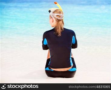 Back side of calm diver girl sitting on seashore, wearing snorkeling equipment and resting on the beach, summer adventure and traveling concept