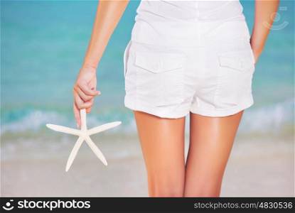 Back side of girl standing on the beach, wearing white beach shorts and holding in hand starfish, summer vacation concept