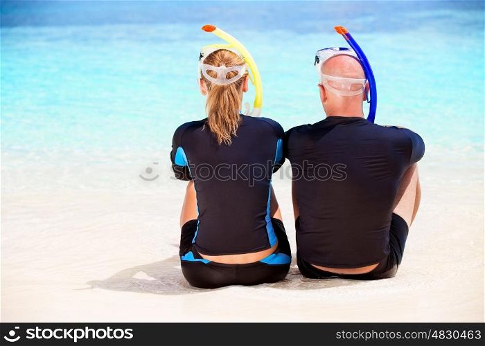 Back side of happy diver couple sitting on the beach and enjoying beautiful sea view, wearing diving equipment and preparing to snorkeling, active summer vacation