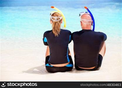 Back side of happy diver couple sitting on the beach and enjoying beautiful sea view, wearing diving equipment and preparing to snorkeling, active summer vacation
