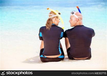 Back side of happy diver couple sitting on the beach and looking on each other with love, wearing diving equipment and preparing to snorkeling, enjoying extreme sport and active summer vacation
