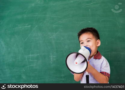 Back to School. Asian funny cute little child boy kindergarten preschool in student uniform speaking through megaphone against on green school blackboard, First time to school education concept