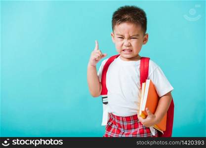 Back to school concept. Portrait Asian cute little child boy face serious hug books thinking and point finger up space, isolated blue background. Kid from preschool kindergarten with school bag