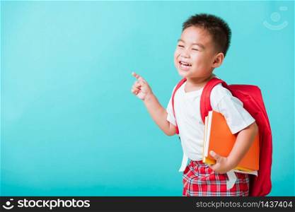 Back to school concept. Portrait Asian happy funny cute little child boy smile hug books and point finger to side away space, isolated blue background. Kid from preschool kindergarten with school bag
