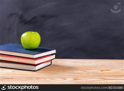 Back to school concept with green apple and books on desk in front of erased black chalkboard.