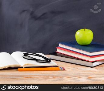 Back to school concept with reading glasses, pencils, notepad, books and green apple on desktop with erased black chalkboard in background.