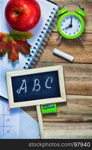Back to School Conceptl: Alarm Clock, Calculator, Blackboard with Letters A, B, C, Pen, Pencils, a Piece of Chalk, Red Apple and Mapple Leaf on the Wooden Background