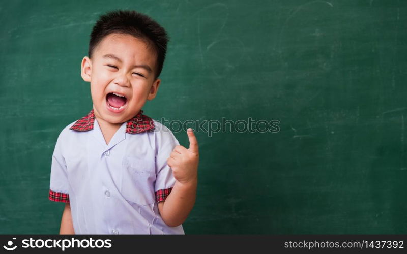 Back to School. Happy Asian funny cute little child boy from kindergarten in student uniform smiling point finger to side away space on green school blackboard, First time to school education concept