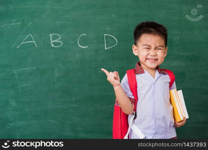 Back to School. Happy Asian funny cute little child boy kindergarten in student uniform and school bag and books smile point finger space side away on school blackboard, First time to school education