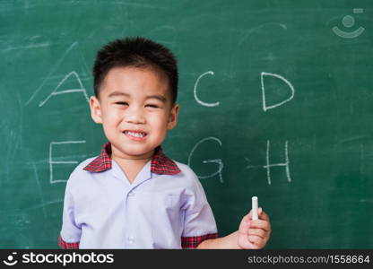 Back to School. Happy Asian funny cute little child boy kindergarten preschool smile in student uniform hold white chalk after write ABC with on green school blackboard, First time to school education