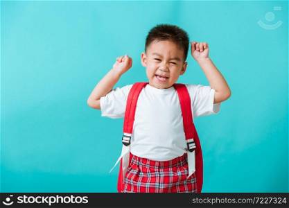 Back to school. Portrait happy Asian cute little child boy in uniform smile raise hands up glad when go back to school, isolated blue background. Kid from preschool kindergarten with school backpack