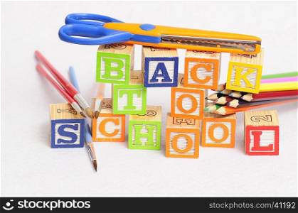 Back to school spelled with alphabet blocks displayed with coloring pencils, paintbrushes and a scissor on a white background