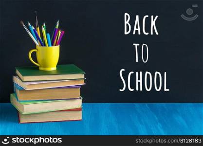 Back to school text on black chalkboard and stationery in a yellow mug on stack of books on blue table. Concept education. Front view.. Back to school text on black chalkboard and stationery in a yellow mug on stack of books on blue table. Concept education. Front view