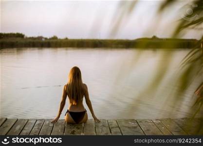 Back view at attractive young woman in bikini sitting on a pier at the lake