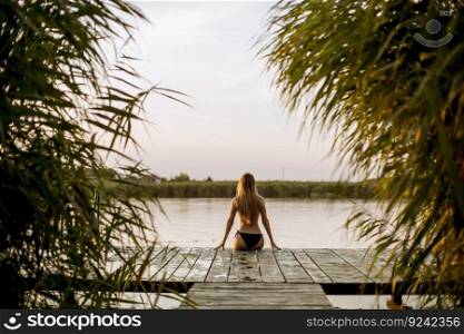 Back view at attractive young woman in bikini sitting on a pier at the lake