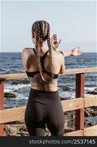 back view athletic woman outdoors by beach stretching while admiring view