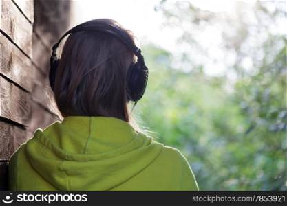 Back view of a girl in headphones listening to music outdoor. She standing by the wooden wall and enjoying nature scene