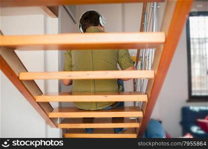 Back view of a man sitting on wooden stairs and enjoying favorite music in wireless headphones. Leisure time and relaxing at home. He likes listening to music sitting on the stairs