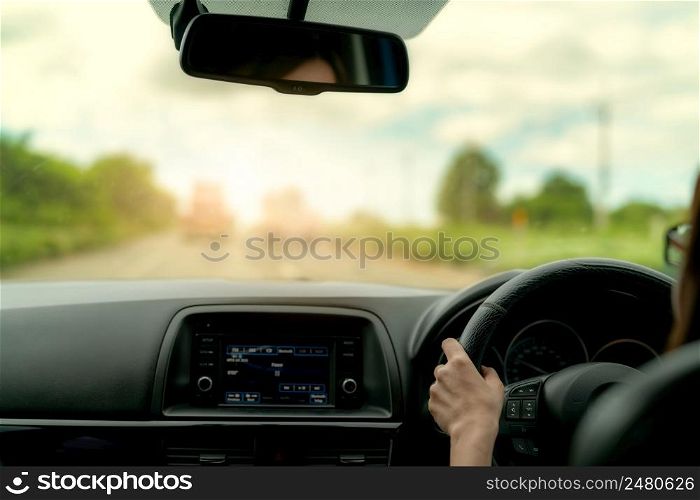 Back view of a woman driving car for summer road trip travel. Car driving with safety on asphalt road. Driver hand holding steering wheel for control car. Inside view of car. Dashboard and windshield.