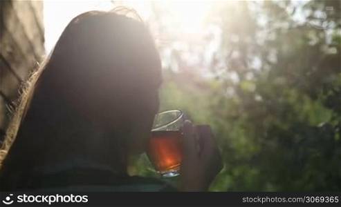 Back view of a woman having tea outdoor during sunset. Evening sun shining through the tree branches
