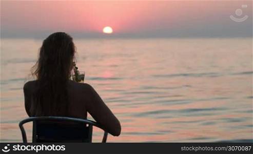 Back view of a woman sitting on the chair with cocktail and enjoying beautiful sunset over the sea