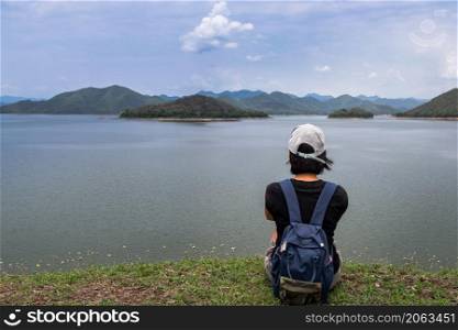 Back view of a young asia woman sitting and enjoying peaceful moment of beautiful view at lake shore with mountains range in background. Holiday travel concept, Rest and enjoyment, lifestyle, Selective focus.
