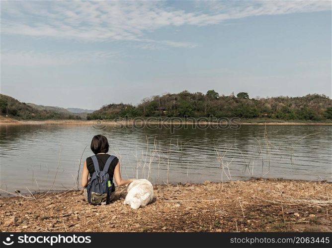 Back view of a young asia woman with white dog sitting and enjoying peaceful moment of beautiful view at lake shore with mountains range in background. Pet and woman, Rest and enjoyment, lifestyle, No focus, specifically.