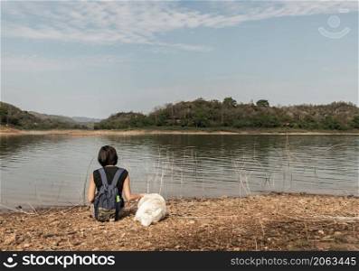 Back view of a young asia woman with white dog sitting and enjoying peaceful moment of beautiful view at lake shore with mountains range in background. Pet and woman, Rest and enjoyment, lifestyle, No focus, specifically.